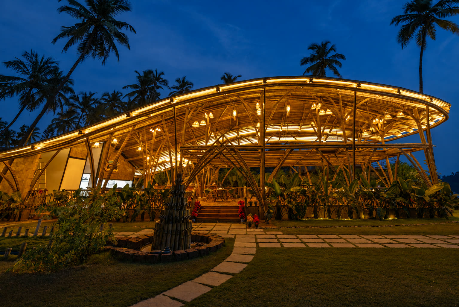 The illuminated bamboo dining room at The Wave Goa beneath palms at blue hour